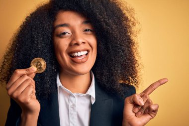 Afro saçlı, sarı arka planda bitcoin tutan Afro-Amerikalı genç bir iş kadını. Elleri ve parmakları kenarda mutlu bir şekilde işaret ediyor.