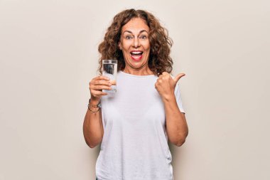 Orta yaşlı, güzel bir kadın beyaz arka planda serinlemek için bir bardak su içiyor baş parmağını yukarı kaldırıyor ve ağzı açık gülümsüyor.