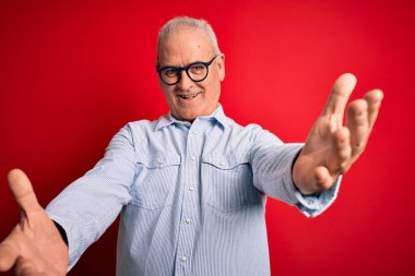 Orta yaşlı, beyaz, sıradan çizgili gömlek ve kırmızı arka planda gözlük takan yakışıklı adam kameraya sarılıp gülümsüyor. Mutluluğu kucaklayan neşeli bir ifade..