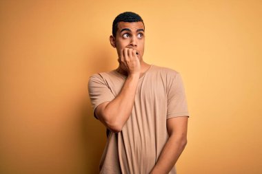 Sarı arka plan üzerinde sıradan tişörtler giyen genç ve yakışıklı Afro-Amerikan erkeği elleri ağız yiyen tırnaklarla gergin ve stresli görünüyor. Endişe sorunu.