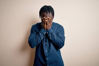 Genç, yakışıklı, Afro-Amerikalı bir adam beyaz arka planda dikilmiş ağlarken suratını ellerle kapatan sıradan bir tişört giyiyor. Depresyon kavramı.