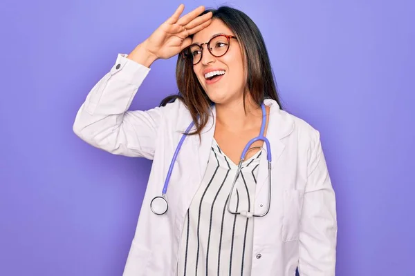 紫色の背景に聴診器や医療用コートを身に着けているプロの医師の女性は非常に満足し 頭の上に手で遠く見て笑顔 検索の概念 — ストック写真