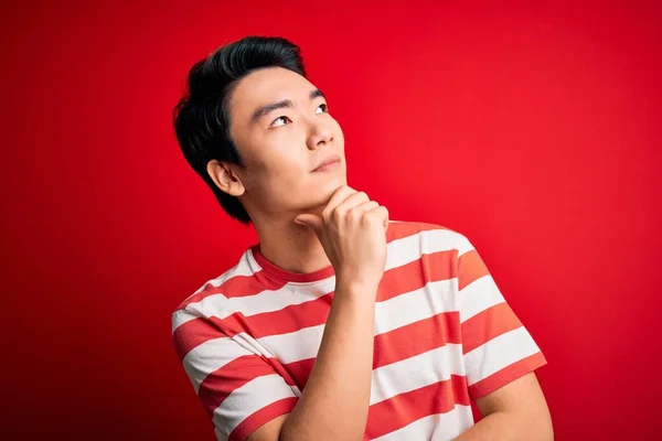 若いハンサムな中国人男性のカジュアルな縞模様のTシャツを着て質問について考えてあごの手で赤い背景の上に立って 集中的な表現 思慮深い顔で微笑んだ 疑いの概念 — ストック写真