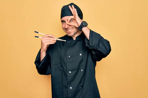 Young handsome cooker man with blue eyes wearing uniform and hat using chopsticks with happy face smiling doing ok sign with hand on eye looking through fingers