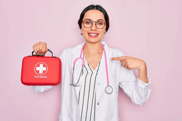 Young beautiful doctor woman with blue eyes wearing stethoscope holding first aid kit box with surprise face pointing finger to himself