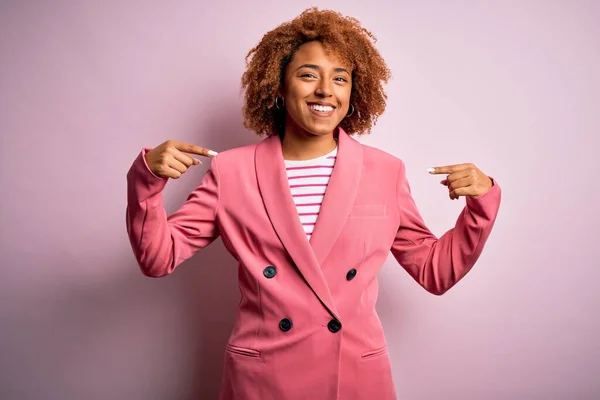 顔に笑顔で自信を持って見えるエレガントなピンクのジャケットを身に着けている巻き毛を持つ若い美しいアフリカ系アメリカ人のビジネス女性 誇りと幸せ指で自分自身を指して — ストック写真