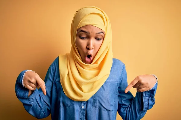 年轻美丽的女人 头戴阿拉伯式传统头巾 头戴黄色背景 手指垂下 露出广告 惊讶的脸和张开的嘴 — 图库照片