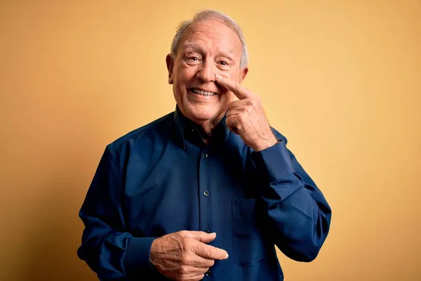 Grey haired senior man wearing casual blue shirt standing over yellow background Pointing with hand finger to face and nose, smiling cheerful. Beauty concept