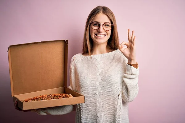 美しい赤頭の女性を保持していますデリバリーボックスでイタリアの新鮮なピザ以上ピンクの背景を持っています指でOkサインをします 優れたシンボル — ストック写真