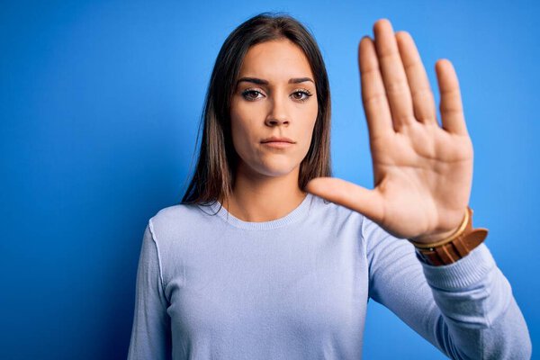 Young beautiful brunette woman wearing casual sweater standing over blue background doing stop sing with palm of the hand. Warning expression with negative and serious gesture on the face.