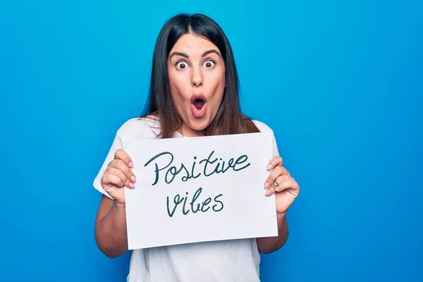 Young beautiful woman asking for optimist attitude holding paper with positive vibes message scared and amazed with open mouth for surprise, disbelief face