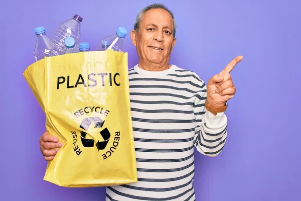 サイドに手と指で非常に幸せなポインティング紫色の背景にリサイクルするプラスチックボトルとシニア男のリサイクルバッグ — ストック写真