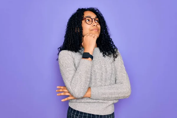 年轻的非洲裔美国女人穿着宽松的毛衣 戴着紫色背景的眼镜 手放在下巴上思考着问题和沉思的表情 面带微笑和体贴的脸怀疑的概念 — 图库照片