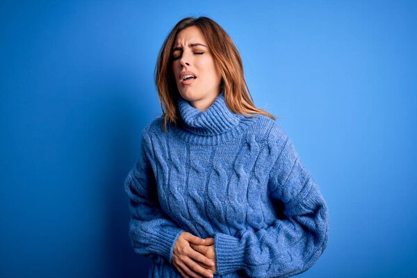 Young beautiful brunette woman wearing casual turtleneck sweater over blue background with hand on stomach because nausea, painful disease feeling unwell. Ache concept.