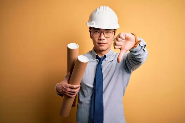 若いハンサムな中国の建築家の男は 怒りの顔を持つ設計図を保持眼鏡と安全ヘルメットを身に着けています 親指ダウンで嫌いを示す負の記号 拒絶コンセプト — ストック写真