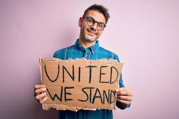 Young handsome activist man asking for union holding banner with united stand message with a happy face standing and smiling with a confident smile showing teeth