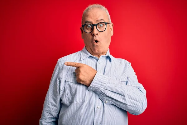 Middle age handsome hoary man wearing casual striped shirt and glasses over red background Surprised pointing with finger to the side, open mouth amazed expression.