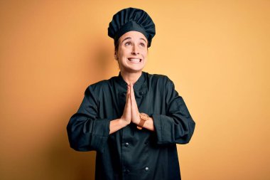 Aşçı üniforması ve şapka giyen genç, güzel bir kadın. Sarı arka planda duruyor. Elleri bağlı yalvarıyor ve dua ediyor. Yalvarıyorum..