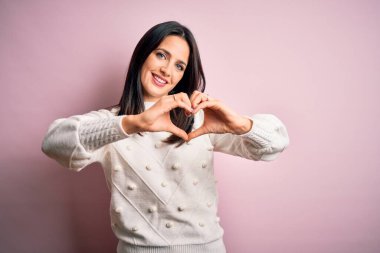 Mavi gözlü genç esmer kadın izole edilmiş pembe arka planda günlük süveter giyiyor ve elleriyle kalp sembolü yapan aşkla gülümsüyor. Romantik konsept.