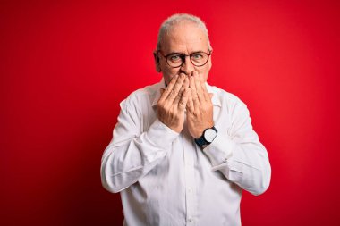 Orta yaşlı yakışıklı, beyaz gömlekli, kırmızı arka planda gözlüklü, gülen, kıkırdayan, ağzı ellerle, dedikoduyla ve skandalla kaplayan bir adam.
