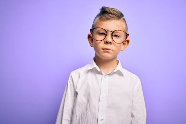 Mavi gözlü, mor arka planda beyaz tişörtlü, yüzünde ciddi bir ifade olan küçük beyaz bir çocuk. Basit ve doğal bir şekilde kameraya bakıyorsun..