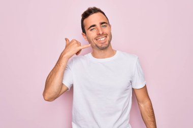 Mavi gözlü yakışıklı bir adam pembe arka planda dikilmiş günlük beyaz tişört giyiyor telefonla el ve parmaklarıyla telefonla konuşuyor gibi gülümsüyor. İletişim kavramları.