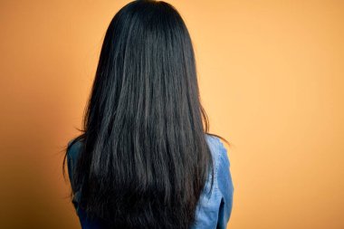 Genç, güzel, Çinli bir kadın, sarı arka planda yalıtılmış, sıradan bir kot gömlek giyiyor ve kollarını kavuşturup arkasına bakıyor.