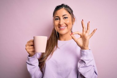 Genç, güzel esmer kadın izole edilmiş pembe arka planda bir kupa kahve içiyor. Tamam işareti yapıyor, mükemmel bir sembol.