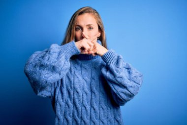 Young beautiful blonde woman wearing casual turtleneck sweater over blue background Rejection expression crossing fingers doing negative sign clipart