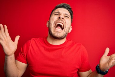 Kırmızı arka plan üzerine günlük tişört giyen mavi gözlü genç adam kızgın ve sinirli bir şekilde bağırıyor ve agresif bir ifade ve kolla bağırıyor. Hayal kırıklığı konsepti.