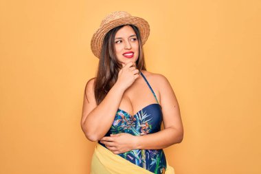 Sarı arka planda mayo ve yaz şapkası takan genç İspanyol kadın. Elleri çenesinde soru sorarken, düşünceli bir ifade takınıyor. Düşünceli bir yüzle gülümsüyor. Şüphe kavramı.