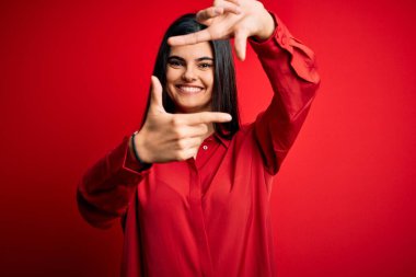 Genç, güzel, esmer, rahat gömlekler giyen, kırmızı arka planda duran, gülümseyen, elleri ve parmakları mutlu yüzlü bir kadın. Yaratıcılık ve fotoğrafçılık kavramı.