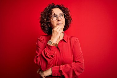 Orta yaşlı güzel kıvırcık saçlı kadın, kırmızı arka planda gözlük takıyor, elleri çenesinde soru sorarken, düşünceli bir ifade takınıyor. Düşünceli bir yüzle gülümsüyor. Şüphe kavramı.