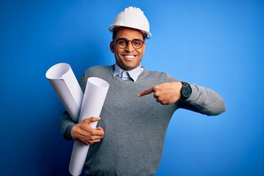 Genç Afrikalı Amerikalı mimar kaskını takmış elinde sürpriz yüzüyle kendini gösteren planlar tutuyor.