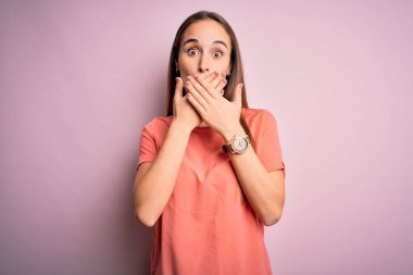 Izole pembe arka plan üzerinde duran rahat t-shirt giyen genç güzel kadın hata için elleri ile ağız kapsayan şok. Gizli kavram.
