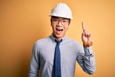 Güvenlik kaskı takan ve sarı arka plan üzerine kravat takan genç, yakışıklı Çinli mimar başarılı bir fikirle işaret ediyor. Heyecanlı ve mutlu. Bir numara..