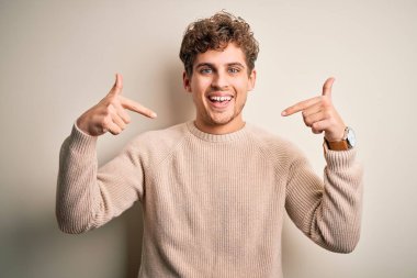 Sarışın, kıvırcık saçlı, beyaz arka planda sıradan süveter giyen, kendine güvenen, kendini gururlu ve mutlu gösteren genç bir adam..