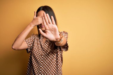 Genç, güzel, esmer bir kadın, izole edilmiş sarı arka plan üzerine gündelik tişört giyiyor, gözleri ellerle örtüyor ve üzgün ve korku ifadeleriyle jest yapıyor. Utanç verici ve olumsuz bir kavram..