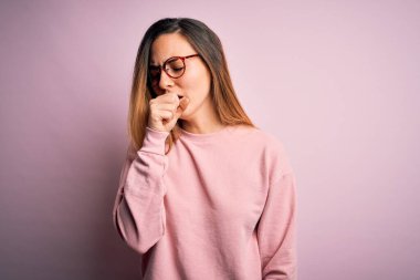 Mavi gözlü, pembe arka planda süveter ve gözlük takan güzel sarışın kadın kendini iyi hissetmiyor ve soğuk algınlığı veya bronşitin belirtisi olarak öksürüyor. Sağlık hizmeti kavramı.