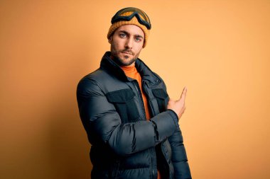 Kar giysileri ve kayak gözlüğü takan sakallı genç yakışıklı kayakçı el parmağı ile reklamını yapıyor, ciddi ve sakin bir yüz ifadesi var.