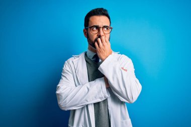 Mavi arka planda ceket ve gözlük takan sakallı yakışıklı doktor elleri ağız yiyen tırnaklarla gergin ve stresli görünüyor. Endişe sorunu.