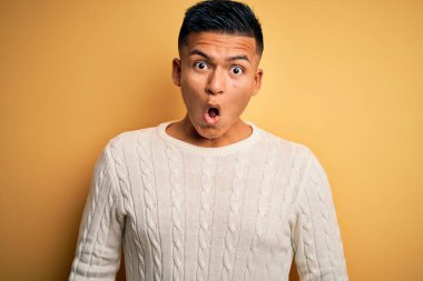 Sarı arka plan üzerine beyaz süveter giyen genç, yakışıklı Latin adam korku ve şaşkınlık dolu bir ifadeyle korku içinde ve şoke olmuştu..