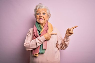 Son sınıf güzeli gri saçlı kadın pembe arka planda günlük süveter ve eşarp giyiyor gülümsüyor ve kameraya bakıyor iki eliyle ve parmaklarıyla yüzünü gösteriyor..
