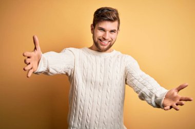 Sarışın, sakallı, mavi gözlü, sarı arka planda beyaz süveter giyen ve kucaklaşmak için kollarını açmış kameraya gülümseyen genç bir adam. Mutluluğu kucaklayan neşeli bir ifade..