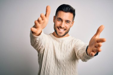 İzole edilmiş beyaz arka plan üzerinde duran sıradan süveter giyen genç yakışıklı adam kucaklanmak için kollarını açmış gülümseyen kameraya bakıyor. Mutluluğu kucaklayan neşeli bir ifade..