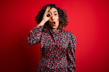 Genç ve güzel kıvırcık Arap kadın. Kırmızı arka planda dikilmiş sıradan bir çiçek elbisesi giyiyor. Şaşırmış bir yüz ve parmaklarından bakan bir gözle şaşırmış bir jest yapıyor. İnançsız ifade.