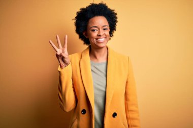 Kıvırcık saçlı, sarı ceketli ve üç numaralı parmaklarını gösterip gülümseyen Afro-Amerikan iş kadını..