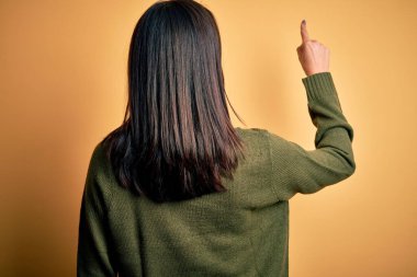 Sarı arka planda yeşil süveter giyen, mavi gözlü, genç esmer kadın, parmak eliyle öne doğru poz veriyor.