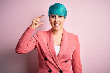 Mavi saçlı, pembe arka planda ceketi olan genç ve güzel bir iş kadını gülümsüyor ve el hareketleriyle küçük beden işareti yaparak kameraya bakıyor. Ölçü kavramı.