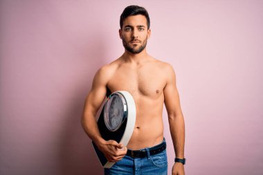 Young handsome slim man with beard shirtless holding weight machine over pink background with a confident expression on smart face thinking serious clipart